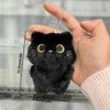 Una mano sostiene un colgante de llavero de un pequeño y esponjoso collar de gato negro contra un fondo desenfocado.