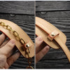A close-up of a tan leather handbag carry loop with a gold chain and a brass clasp being held by a hand.