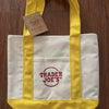 A yellow canvas tote bag with white stripes and a red logo tag resting on a wooden surface.