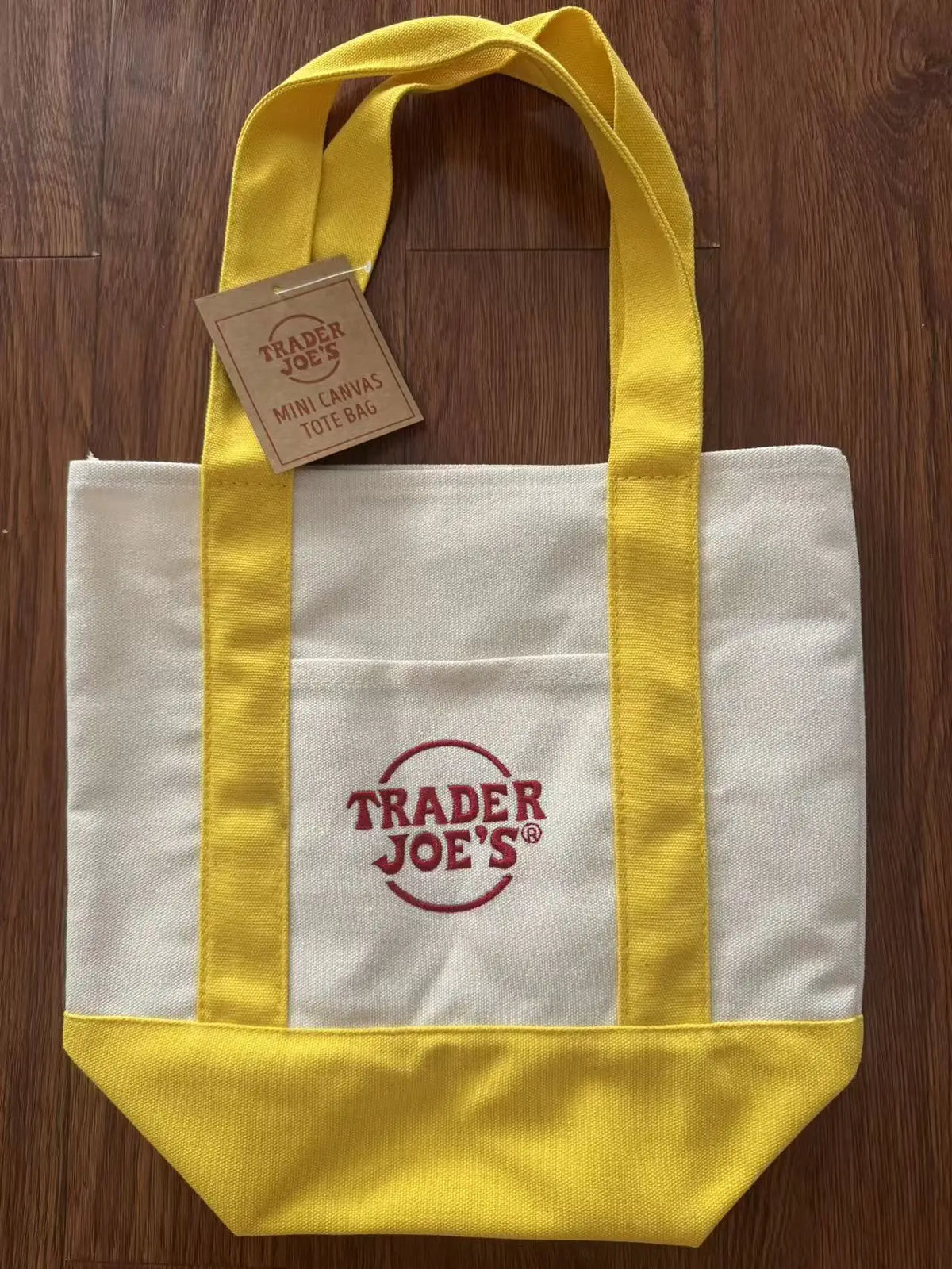 A yellow canvas tote bag with white stripes and a red logo tag resting on a wooden surface.