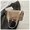A tan vintage curved flap shoulder bag is shown being held by a hand against a dark gray blazer.