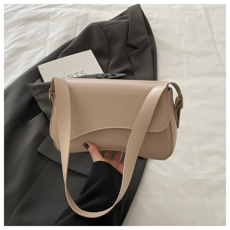 A tan vintage curved flap shoulder bag is shown being held by a hand against a dark gray blazer.