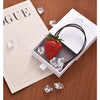 Un charm porte-clés en forme de fraise rouge est exposé sur une boîte blanche avec des glaçons sur une surface en bois.
