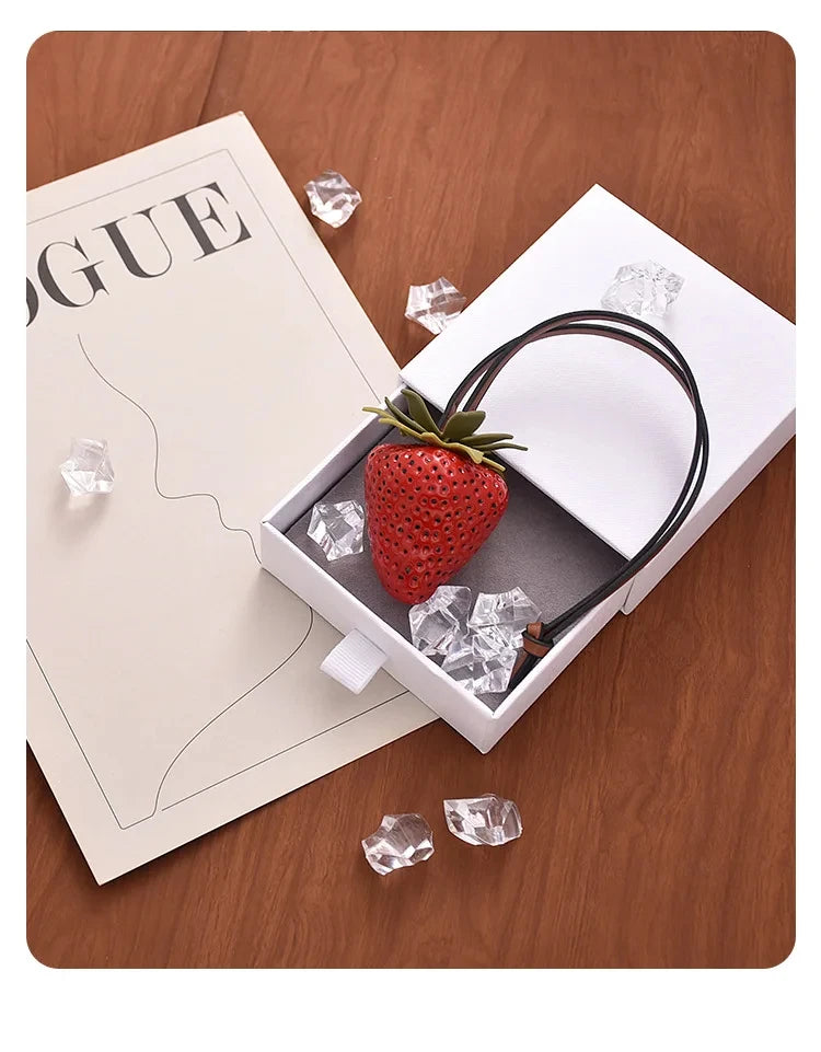 Un charm porte-clés en forme de fraise rouge est exposé sur une boîte blanche avec des glaçons sur une surface en bois.