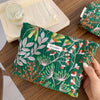 A dark green cosmetic pouch with floral embroidery rests on a wooden table next to an open book.