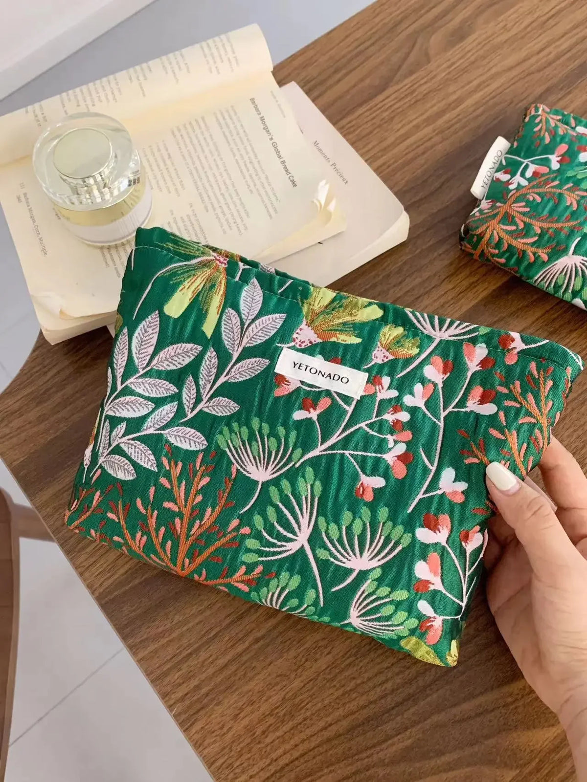 A dark green cosmetic pouch with floral embroidery rests on a wooden table next to an open book.