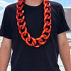 A man wearing a chunky gold acrylic Cuban link necklace with an orange-red color against a black t-shirt.