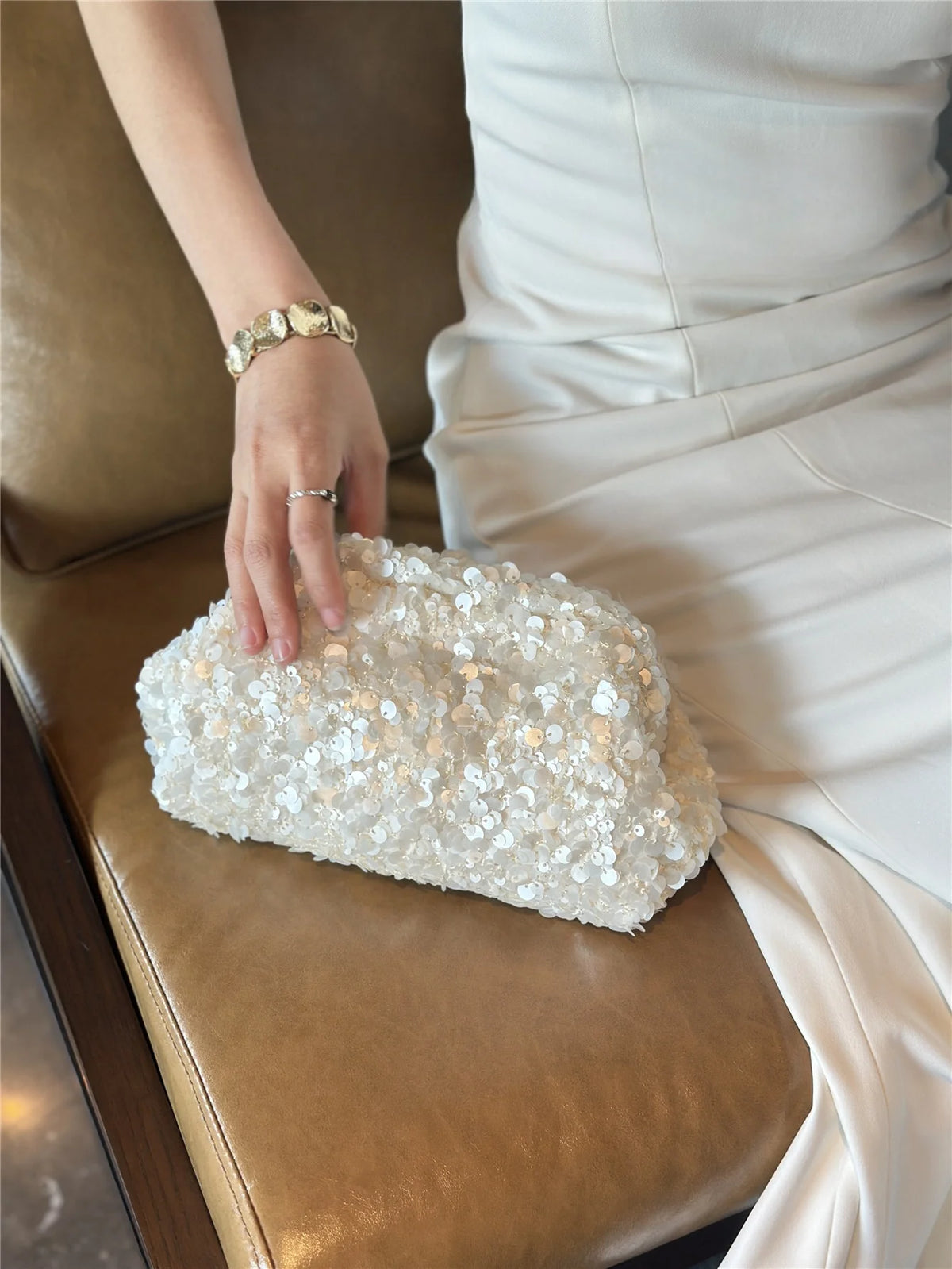 A hand places a white beaded chain shoulder clutch bag on a brown leather sofa.