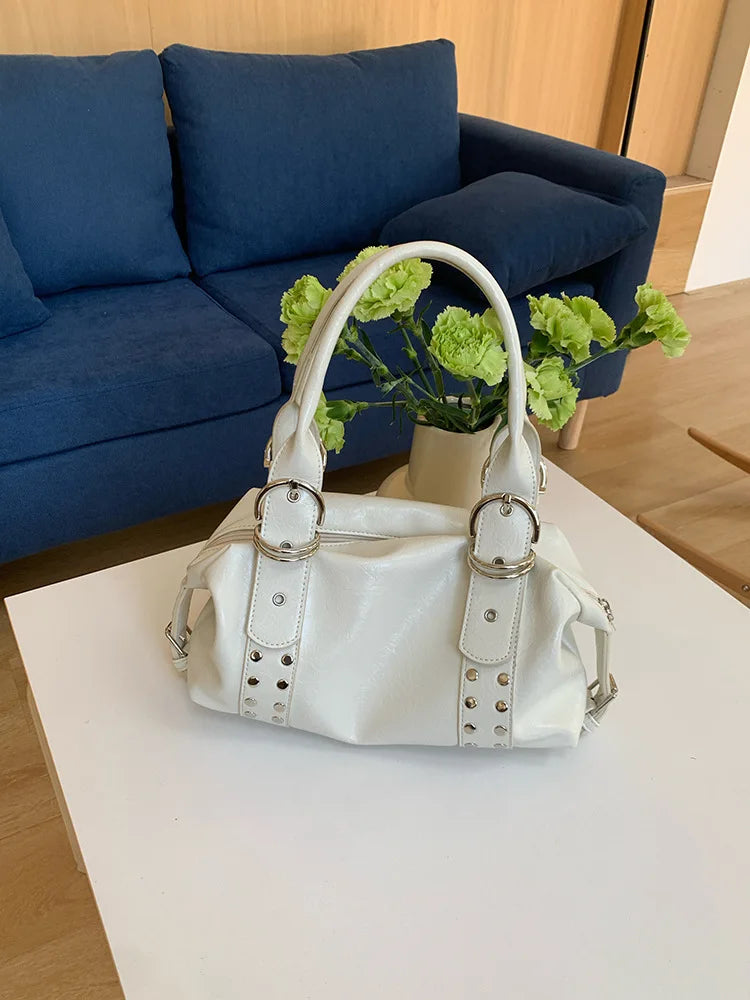 A white PU leather shoulder bag with silver rivets and a handle is displayed on a white table next to a blue sofa.