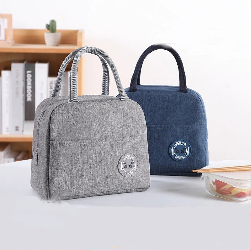 A gray insulated lunch bag with black handles is displayed on a white surface next to a dark blue version.