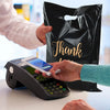 A hand is placing a credit card into a black electronic device next to a black plastic gift bag with gold lettering.