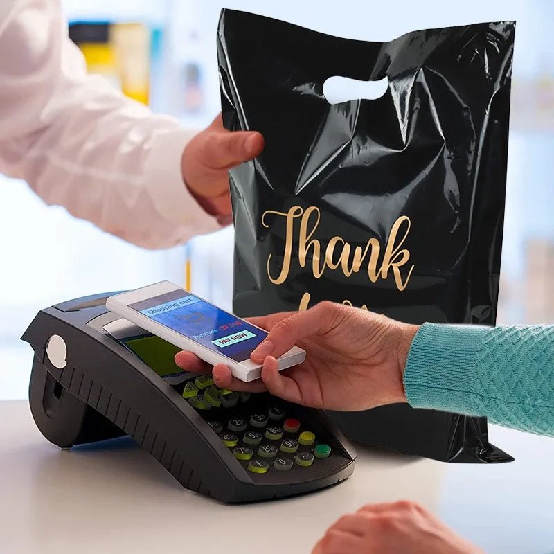 A hand is placing a credit card into a black electronic device next to a black plastic gift bag with gold lettering.