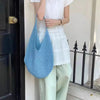 A woman wearing a white top and light green pants stands next to a black door, holding a light blue crochet mesh shoulder bag
