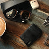 A close-up shot of a black slim minimalist men's wallet with RFID protection resting on a wooden surface next to sunglasses a