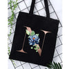 Black canvas tote bag featuring a large gold letter N with blue and green floral accents.