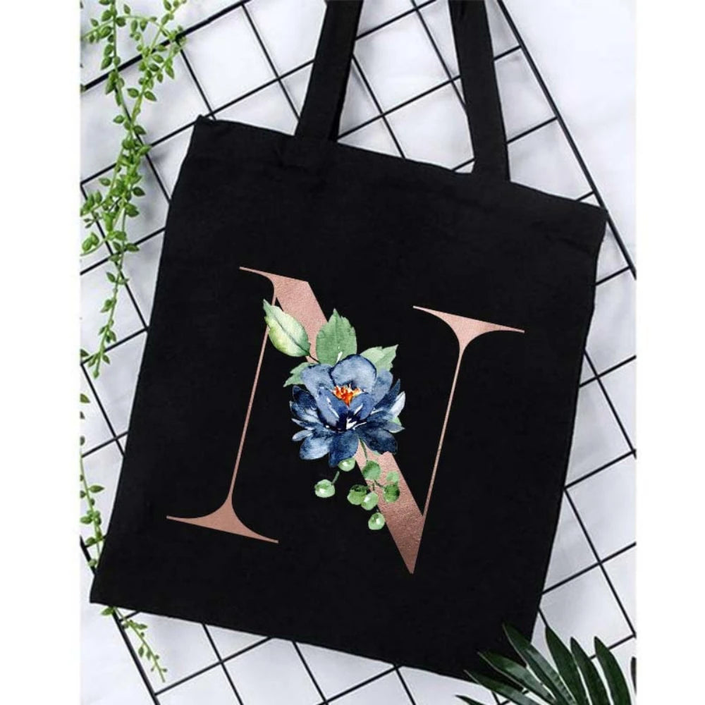 Black canvas tote bag featuring a large gold letter N with blue and green floral accents.