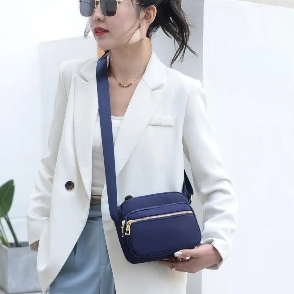 A woman wearing a white blazer and light blue skirt holds a navy blue compact oxford fabric crossbody bag.