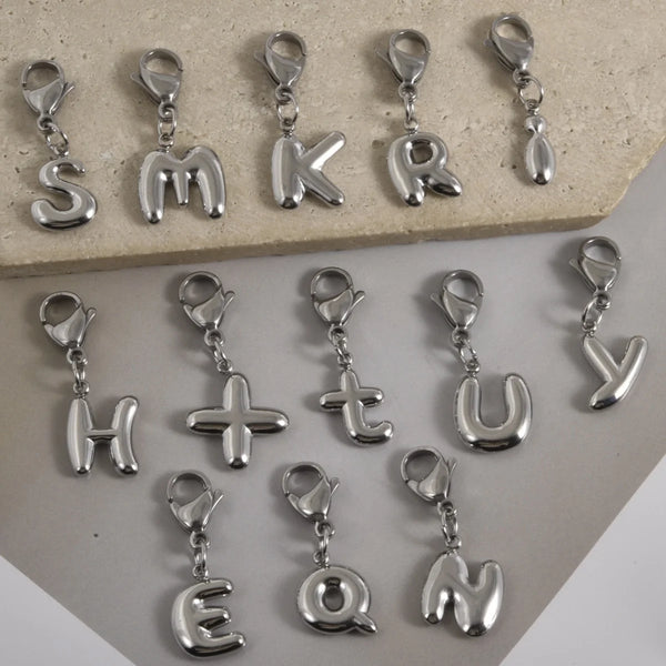 A collection of stainless steel alphabet letter charms with lobster clasps arranged on a light surface.