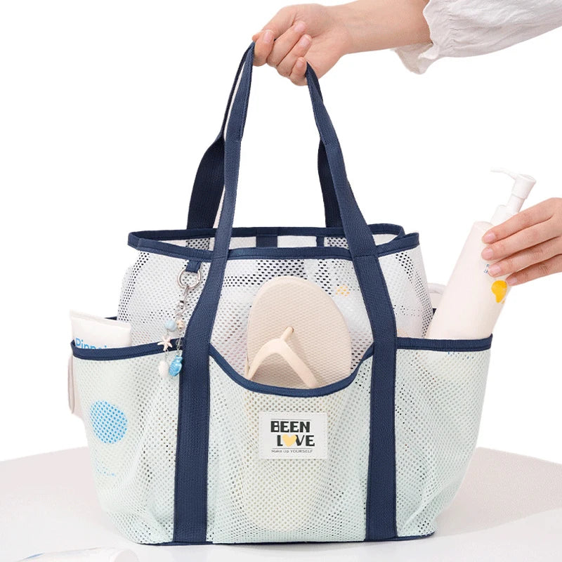 A hand is holding a light blue mesh beach tote bag with dark blue handles and a white bottle.