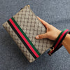Eine Hand hält eine beige-grüne Gucci-Stil-Clutchtasche mit rot-schwarz gestreiftem Lederriemen vor einem grauen Hintergrund
