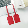 A red PU leather luggage tag with a strap and buckle is shown next to a white laptop and a book.