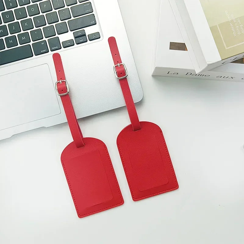 A red PU leather luggage tag with a strap and buckle is shown next to a white laptop and a book.