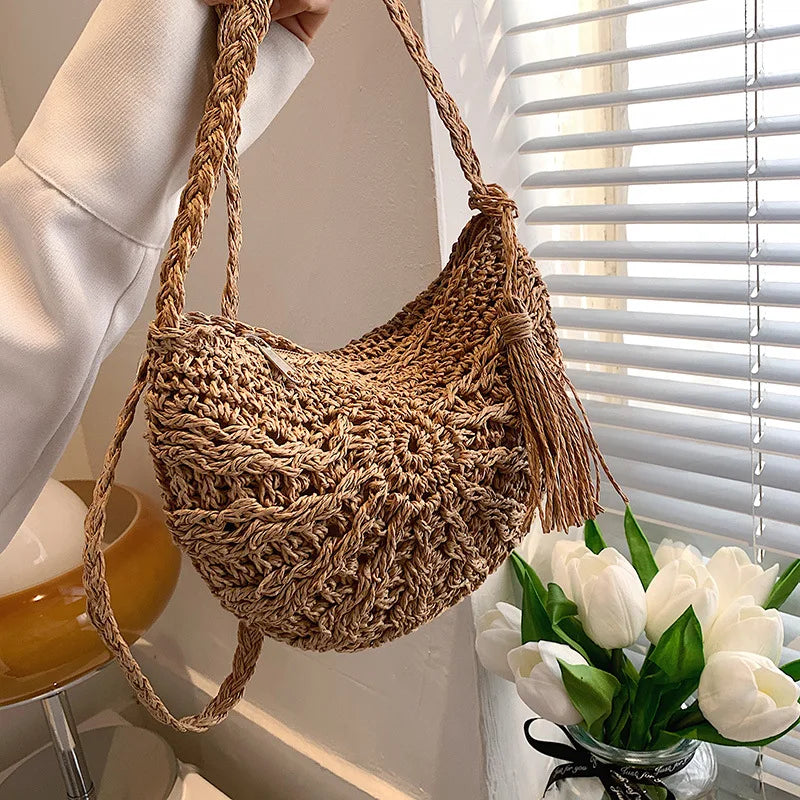 A handmade rattan straw crossbody bag is hanging on a hook next to a window with white blinds.