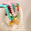 A colorful bead necklace featuring pink, blue, and green resin heart charms displayed on a white ceramic surface.