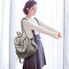 Une femme portant un sac à dos en cuir gris souple et un chemisier blanc se tient devant une fenêtre transparente.