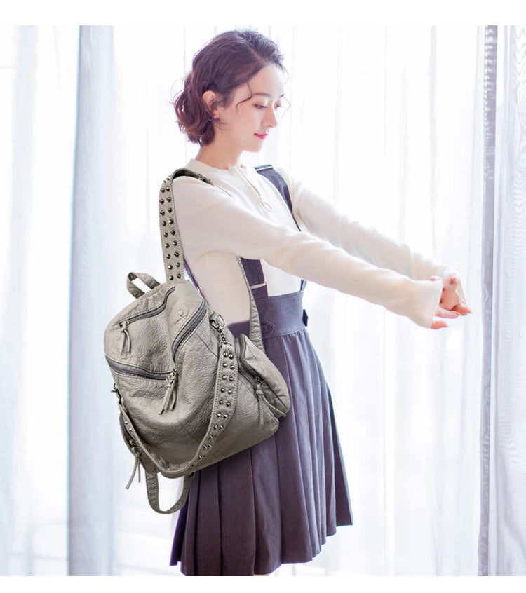 Une femme portant un sac à dos en cuir gris souple et un chemisier blanc se tient devant une fenêtre transparente.