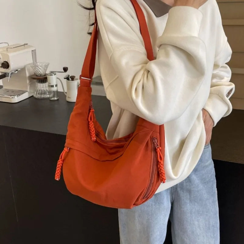 Eine Frau hält eine orangefarbene Nylon-Crossbody-Tasche gegen einen weißen Pullover in einer Küche.