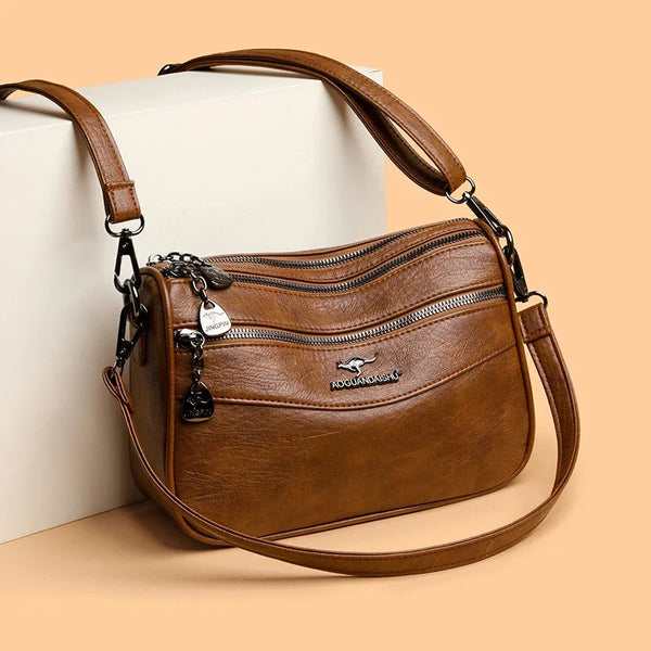 A brown leather trendy women's crossbody bag with gold hardware resting on a light colored box.
