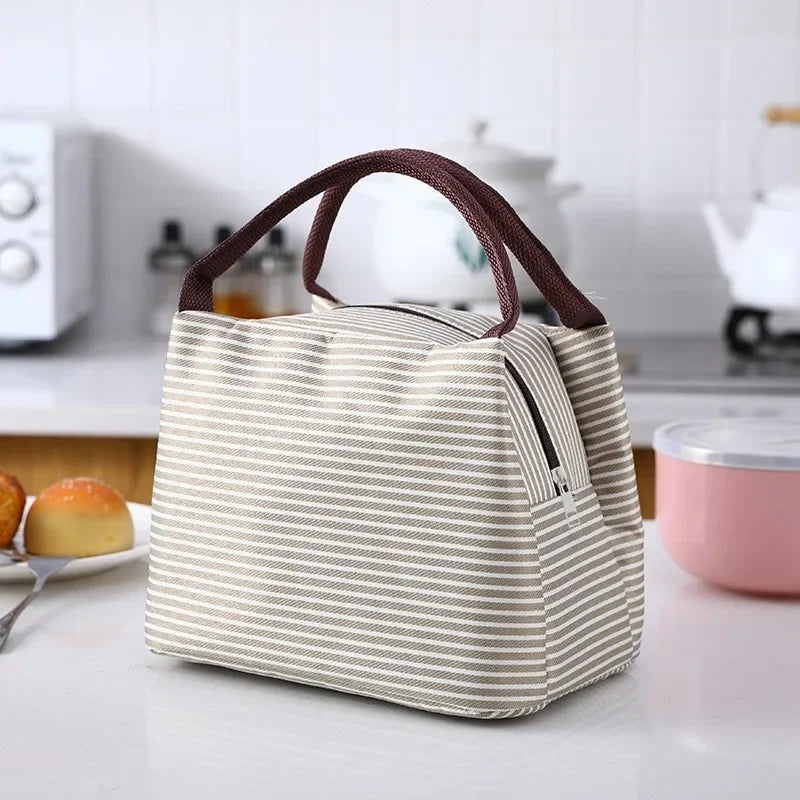 A beige and white striped thermal lunch bag with brown handles resting on a white table.