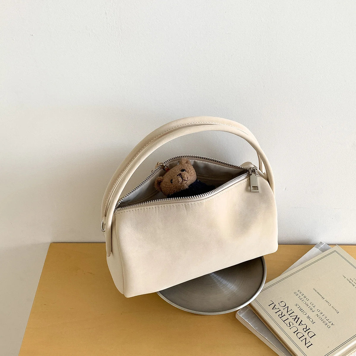 A light beige faux suede crossbody bag with a gold zipper and a brown pom-pom resting inside, placed on a wooden surface.