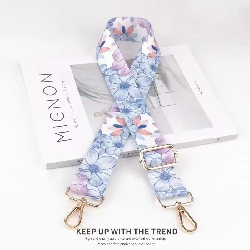 A light blue and purple floral patterned fabric strap with gold hardware displayed on a white background.