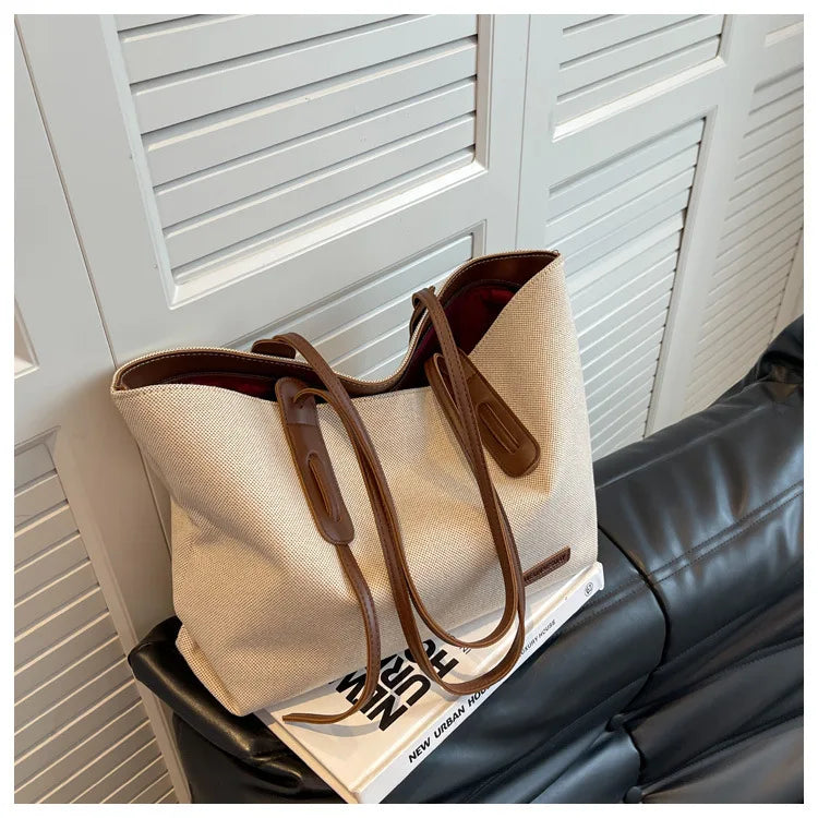 A beige canvas shoulder tote bag with brown leather accents resting on a black leather sofa.