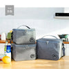 Three grey insulated portable lunch bags with shoulder straps are displayed on a wooden table.