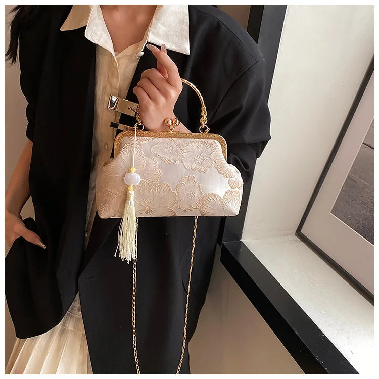 A woman wearing a black blazer and light top holds an embroidered silk evening clutch with a gold chain strap.