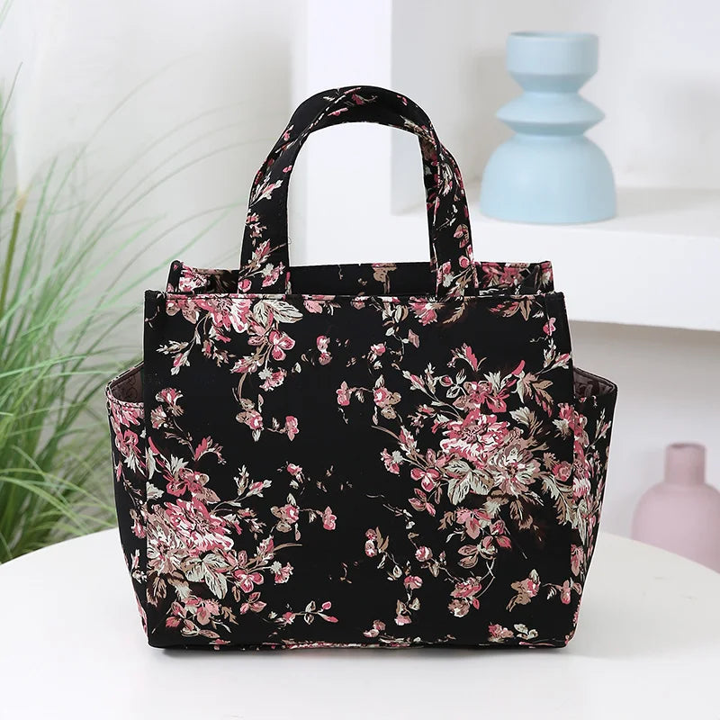 A floral patterned black cloth tote bag with pink and green leaves sitting on a white surface.