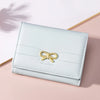 Portefeuille à cartes tri-pli bleu clair en PU avec un nœud tressé or, posé sur une surface rose.