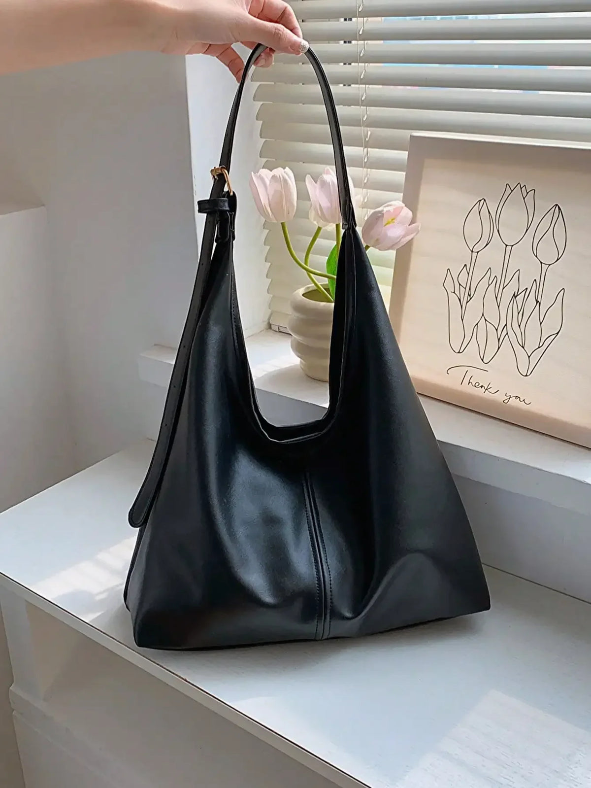 A black PU leather slouchy hobo shoulder bag is displayed on a white shelf next to pink tulips.