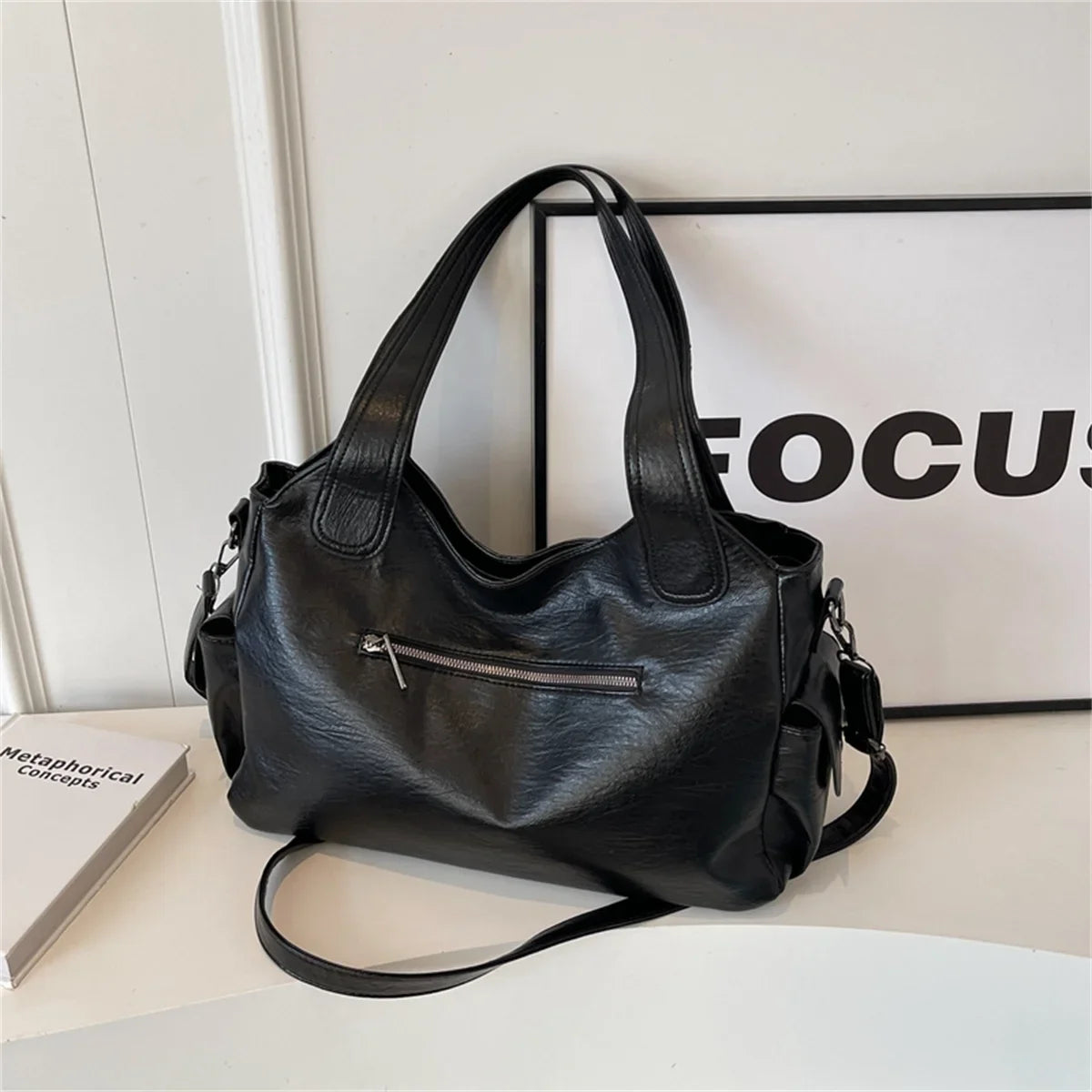 Un sac fourre-tout en cuir noir à fermeture éclair est exposé sur une surface blanche à côté d'un panneau encadré.