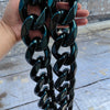 A hand holds a chunky black acrylic Cuban link necklace for men against a light gray outdoor background.