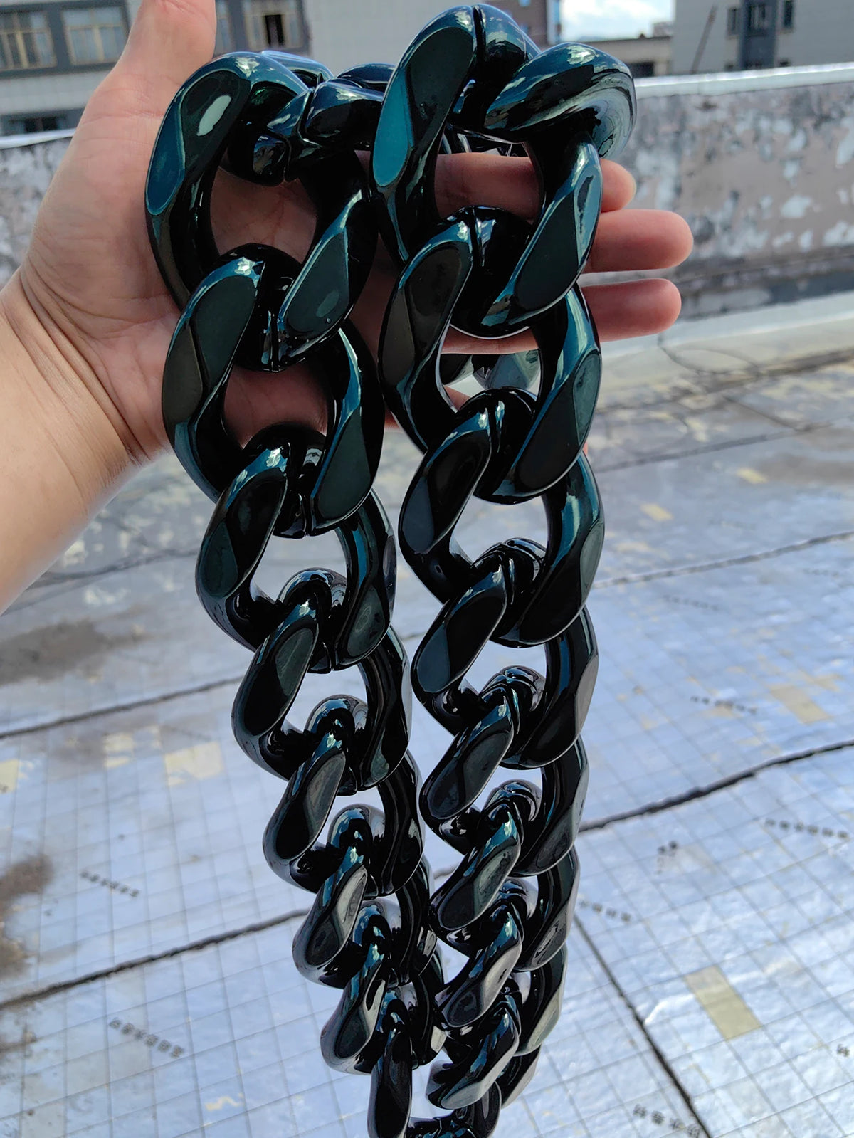 A hand holds a chunky black acrylic Cuban link necklace for men against a light gray outdoor background.