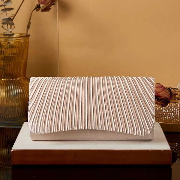 A beige satin evening clutch with vertical pleats rests on a wooden block against a warm brown background.