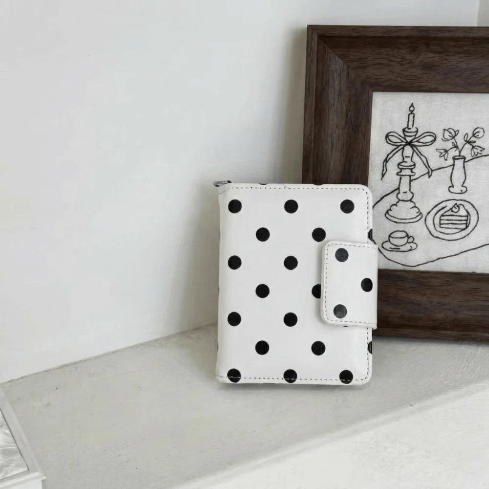 A white polka dot passport holder with multi-card slots resting in front of a dark wooden framed picture.