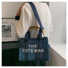 A woman is holding a dark blue canvas tote bag with light brown leather handles and a navy strap, set against a white backgro