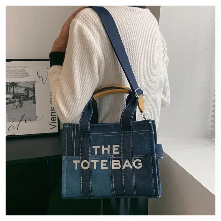A woman is holding a dark blue canvas tote bag with light brown leather handles and a navy strap, set against a white backgro