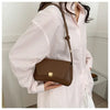 A woman holds a brown leather crossbody bag with a long strap against a pink striped shirt.