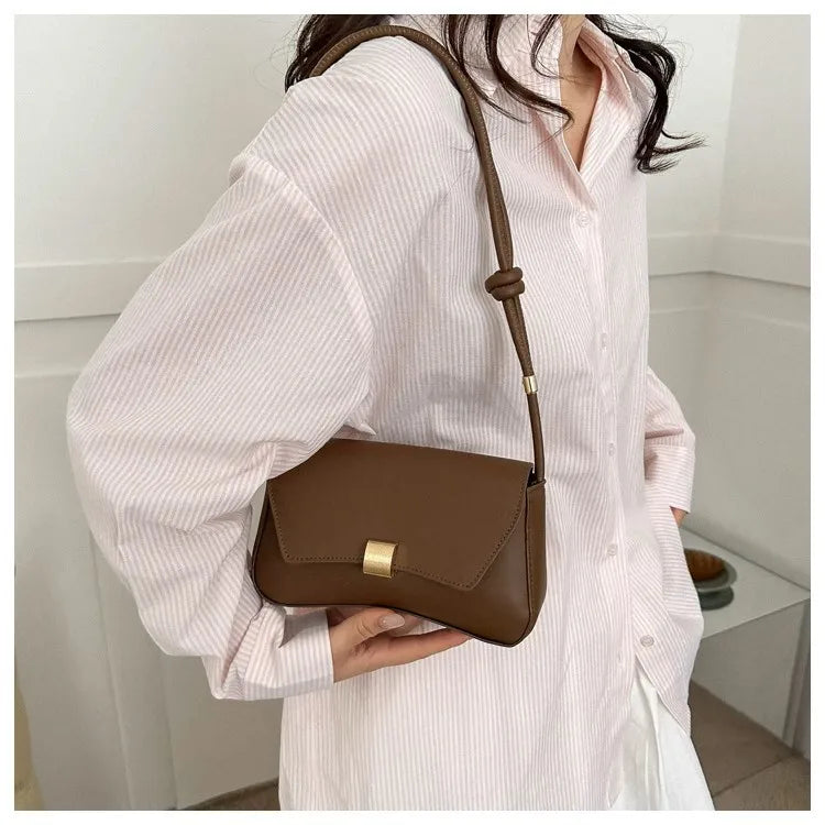 A woman holds a brown leather crossbody bag with a long strap against a pink striped shirt.